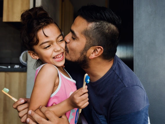 man kisses daughter on cheek while they hold toothbrushes