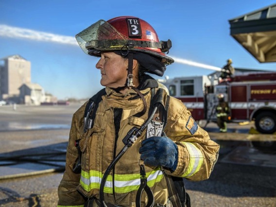 A woman in firefighting gear with a firetruck in the background looks in the direction of a structure that is being sprayed with water during a training exercise. 