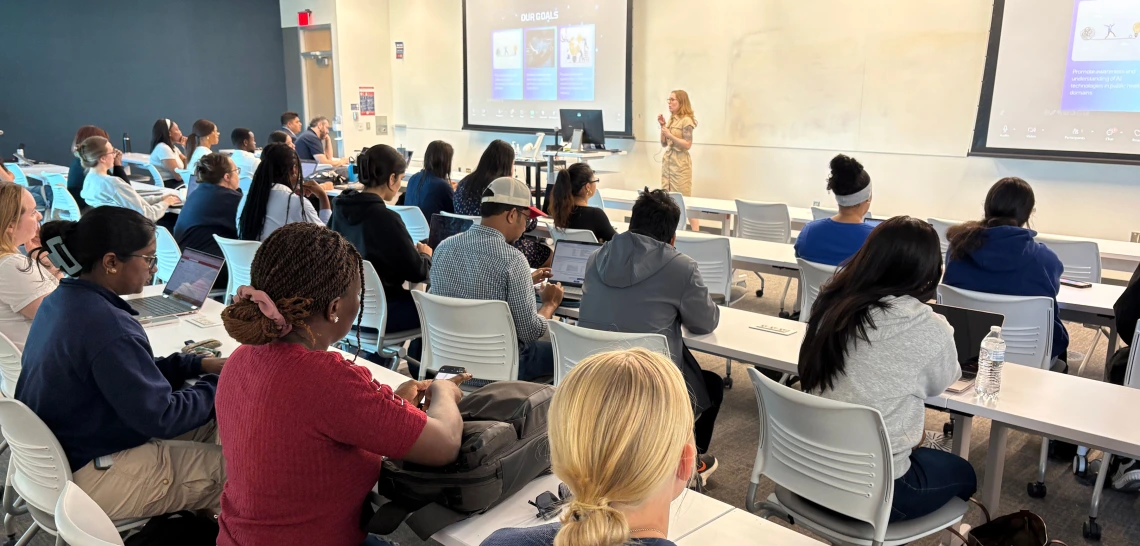Photo of a room full of people sitting at desks while a woman speaks at the front of the room. 