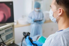 A doctor wearing surgical gear looks at a computer screen as he conducts a colonoscopy on a patient.